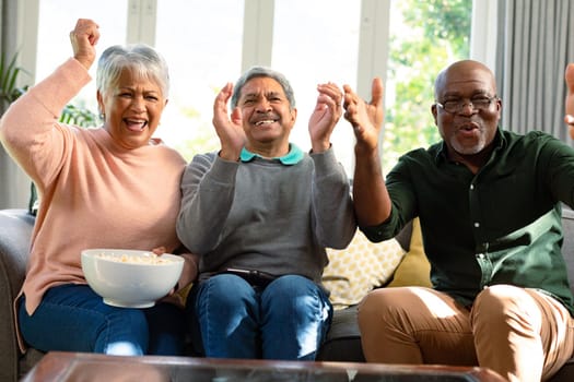 Two happy diverse senior couple and their african american male friend watching tv and having fun. retirement lifestyle relaxing at home with technology.