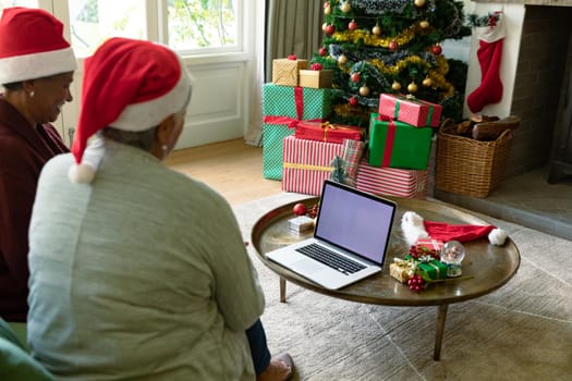 Two diverse senior female friends wearing santa hats using laptop with copy space at christmas time. christmas, festivity and communication technology.