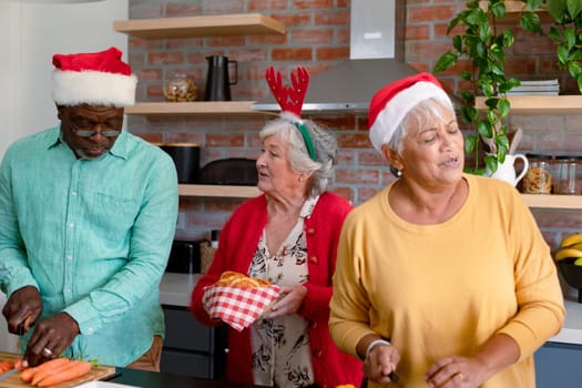 Three diverse senior male and female friends in christmas hats cooking together in kitchen. christmas festivities, celebrating at home with friends.