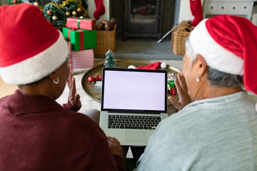 Two diverse senior female friends wearing santa hats using laptop with copy space at christmas time. christmas, festivity and communication technology.