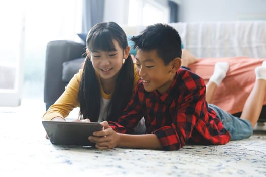 Smiling asian brother and sister lying in living room and using tablet. childhood leisure time at home with technology.