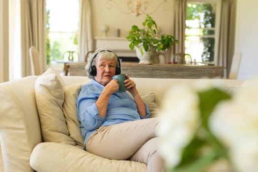 Senior caucasian woman sitting on sofa wearing headphones and holding mug. retreat, retirement and happy senior lifestyle concept.