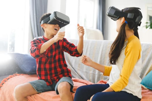 Smiling asian brother and sister sitting on couch and using vr headsets. childhood leisure time and discovery using technology at home.