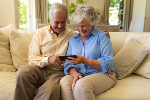 Senior caucasian couple sitting on sofa using smartphone and smiling. retreat, retirement and happy senior lifestyle concept.