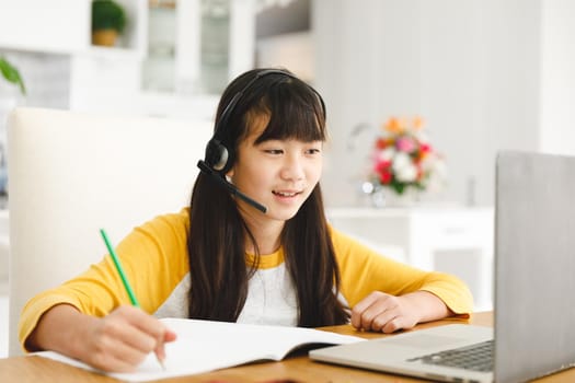 Asian girl sitting at table and using laptop during online lessons. childhood, education and discovery using technology at home.