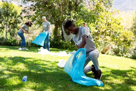 Smiling african american man holding refuse sack, collecting plastic waste in field with friends. eco conservation volunteers doing countryside clean-up.