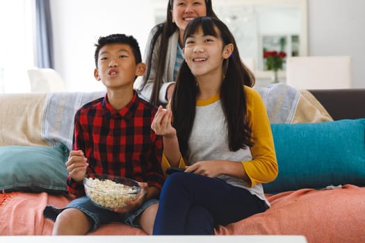 Excited asian mother with son and daughter sitting on couch watching tv and eating popcorn. family entertainment and leisure time together at home.