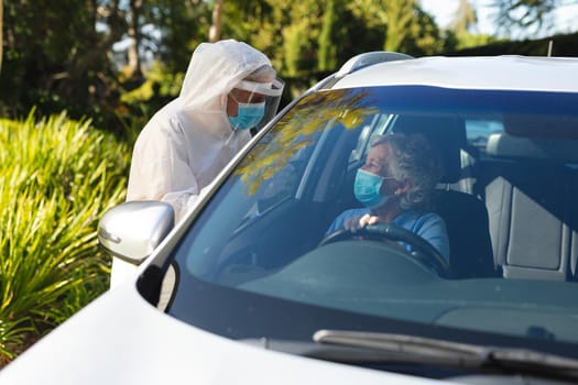 Medical worker wearing ppe suit taking temperature of senior caucasian woman sitting in car. retreat, retirement and senior lifestyle during covid 19 pandemic concept.