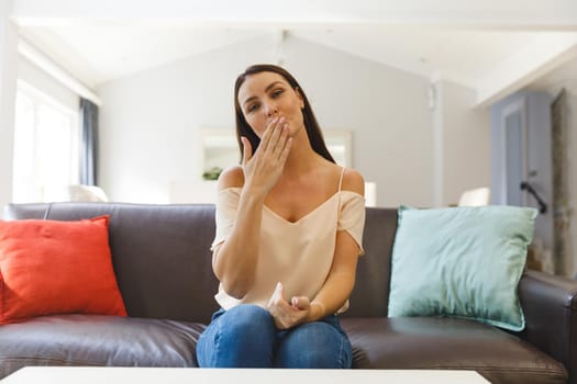 Happy caucasian woman sitting on couch having video call in living room and blowing a kiss. keeping in touch, leisure time at home with communication technology.