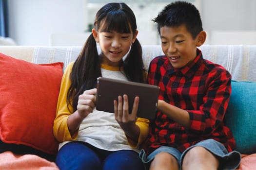 Smiling asian brother and sister sitting on couch and using tablet. childhood leisure time at home with technology.