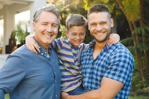 Portrait of smiling caucasian grandfather with adult son and grandson outside house in garden. multi generation family enjoying leisure time together at home.
