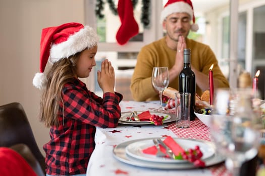 Caucasian daughter and father wearing santa hats praying at christmas table. family christmas time and festivity together at home.