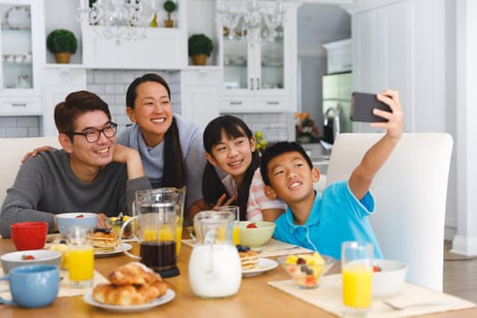 Smiling asian parents with son and daughter sitting at breakfast table taking selfie. family enjoying mealtime together at home.