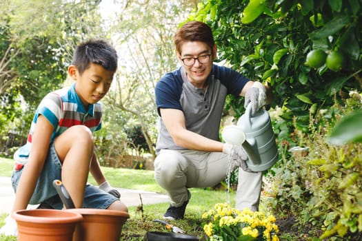 Happy asian father and son smiling, wearing gloves and watering plants together in garden. family leisure time at home gardening.