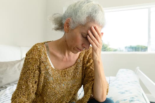 Portrait of tired caucasian senior woman with headache sitting on bed at home. senior health and lifestyle at home.