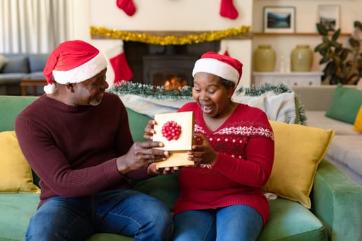 Surprised african american senior couple in santa hats sharing presents at christmas time. retirement lifestyle and christmas festivities, celebrating at home.