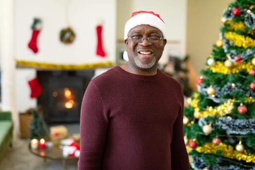 Happy african american senior man in santa hat looking at camera at christmas time. retirement lifestyle and christmas festivities, celebrating at home.