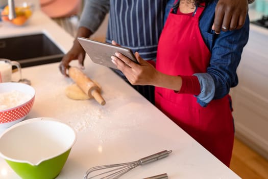 Midsection of african american couple wearing aprons, baking together and using tablet. family time, having fun together at home.