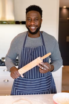 Portrait of happy african american man wearing apron, holding rolling pin in kitchen. cooking and baking, spending time at home.