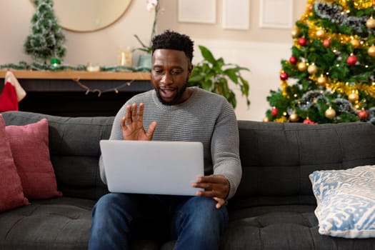 Happy african american man sitting on sofa, having video call, christmas decorations in background. christmas, festivity and communication technology.