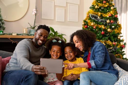 Happy african american family having video call on tablet, christmas decorations in background. christmas, festivity and communication technology.