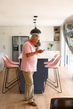 Relaxed caucasian senior man standing in kitchen, drinking coffee and using smartphone. healthy retirement lifestyle at home.