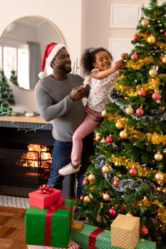 Happy african american father wearing santa hat and daughter decorating christmas tree. christmas, festivity and tradition at home.