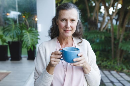 Portrait of happy senior caucasian woman looking to camera, holding cup in garden. retirement lifestyle, spending time alone at home.