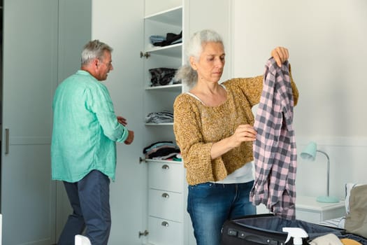 Focused caucasian senior couple packing suitcase together in bedroom. travel preparation during covid 19 pandemic.