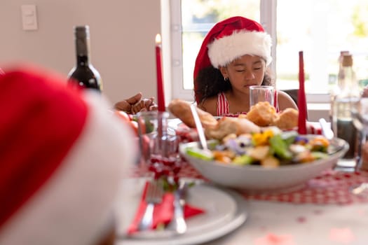 African american girl wearing santa hat praying at christmas table. christmas, festivity and tradition at home.