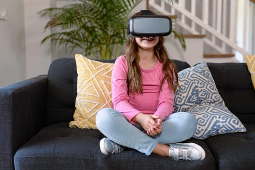 Happy caucasian girl sitting on sofa, wearing vr headset. childhood, leisure and discovery using technology at home.