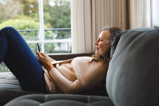 Happy senior caucasian woman in living room sitting on sofa, using smartphone. retirement lifestyle, spending time alone at home with technology.