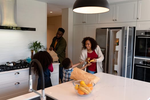 Happy african american siblings unpacking groceries with parents in kitchen. family time, having fun together at home.