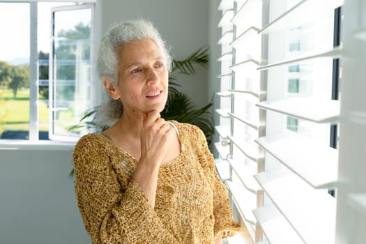 Thoughtful caucasian senior woman standing at window, looking into distance. healthy retirement lifestyle at home.