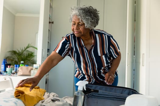 Focused african american senior woman packing suitcase in bedroom. travel preparation during covid 19 pandemic.