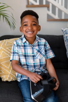 Happy african american boy sitting on sofa and holding vr headset. childhood, leisure and discovery using technology at home.