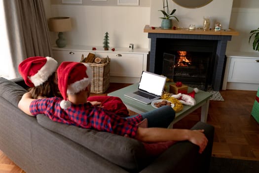 Back view of caucasian couple wearing santa hats, using laptop with copy space at christmas time. christmas, festivity and communication technology.