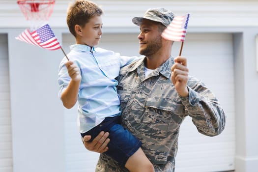 Smiling caucasian male soldier with son outside house holding american flags. soldier returning home to family.
