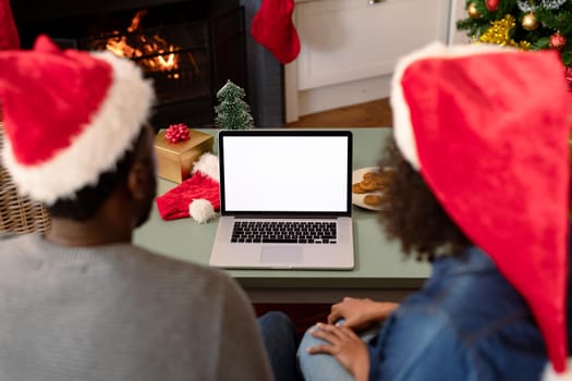 Back view of african american couple wearing santa hats, using laptop with copy space on screen. christmas, festivity and communication technology.