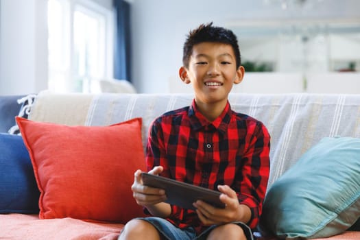 Portrait of smiling asian boy sitting on couch and using tablet. childhood leisure time at home with technology.