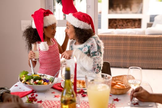 Happy african american mother and daughter wearing santa hats talking at christmas table. family christmas time and festivity together at home.