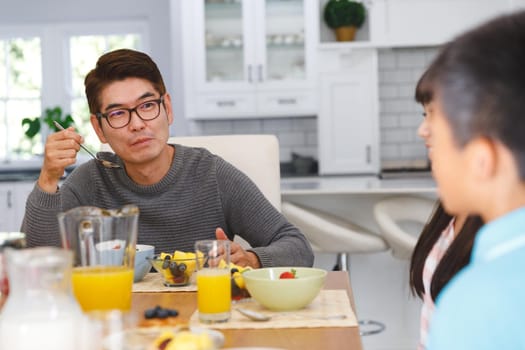 Asian father with son and daughter sitting at table having conversation at breakfast. family enjoying mealtime together at home.