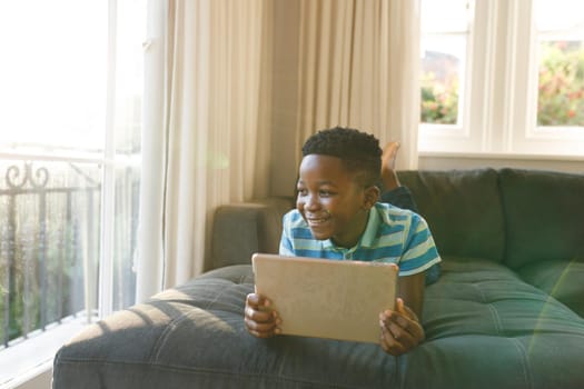 Smiling african american boy using tablet and lying on couch in living room. spending time alone with technology at home.