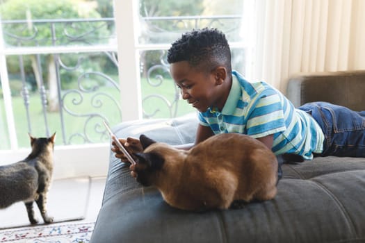Happy african american boy using tablet and lying on couch with cats in living room. spending time alone at home with technology.