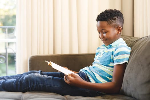 Smiling african american boy reading book and sitting on couch in living room. spending time alone at home.