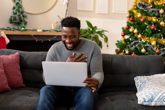 Happy african american man sitting on sofa, having video call, christmas decorations in background. christmas, festivity and communication technology.
