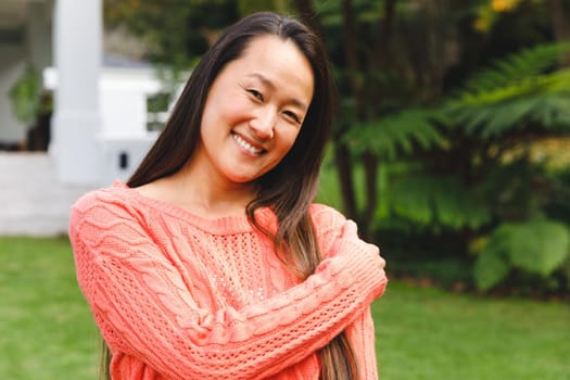 Portrait of happy asian woman smiling in garden outside family home wearing pink sweater. enjoying leisure time alone at home.