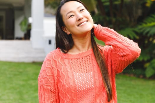 Portrait of happy asian woman smiling in garden outside family home wearing pink sweater. enjoying leisure time alone at home.