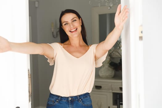 Portrait of happy caucasian woman standing at door and greeting a visitor with open arms. hospitality and welcoming guests at home