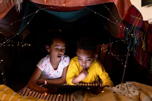 Surprised african american siblings lying in makeshift tent, using tablet. childhood, christmas, festivity and tradition at home.